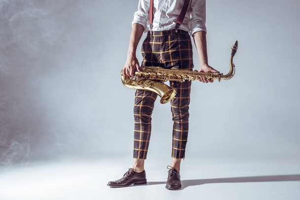 plan recadré de jeune musicien élégant debout et tenant le saxophone en fumée sur gris
 - Photo, image