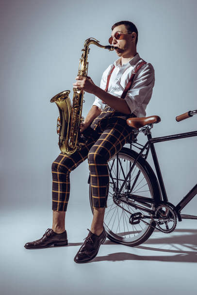 handsome young performer in sunglasses playing saxophone while sitting on bicycle on grey - Photo, Image