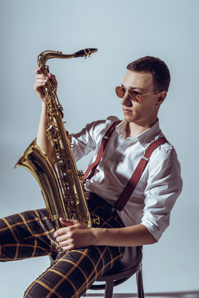 guapo joven artista en gafas de sol sosteniendo saxofón y mirando hacia otro lado en gris
 - Foto, imagen