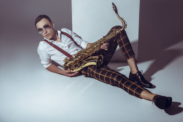 guapo joven músico con estilo en gafas de sol acostado con saxofón en gris
 - Foto, Imagen