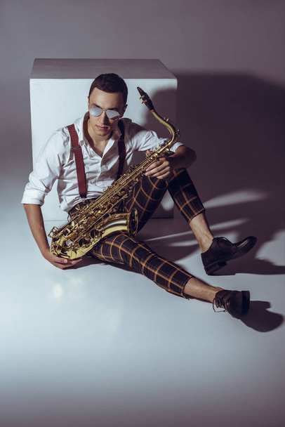 Stilvoller junger Musiker mit Sonnenbrille sitzt mit Saxofon auf grau - Foto, Bild