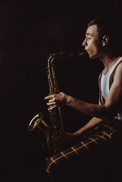 side view of stylish young saxophonist sitting and playing sax on black - Φωτογραφία, εικόνα