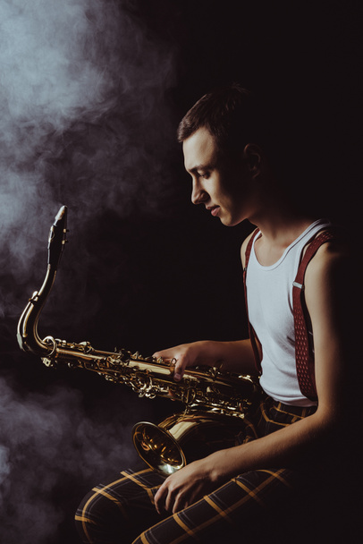 side view of stylish young jazzman sitting and holding saxophone in smoke on black  - Фото, зображення