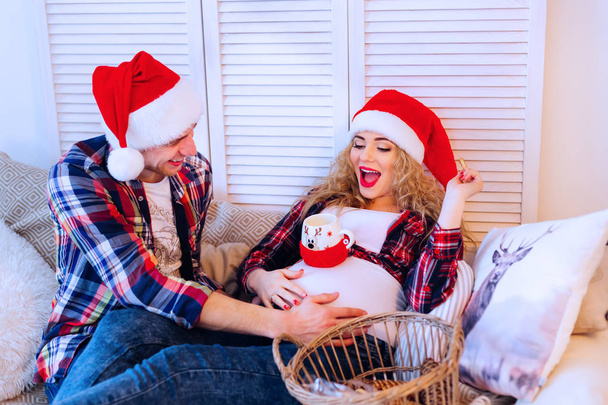 a man in Santa Clause's hat and his pregnant wife sitting on the couch - Photo, Image