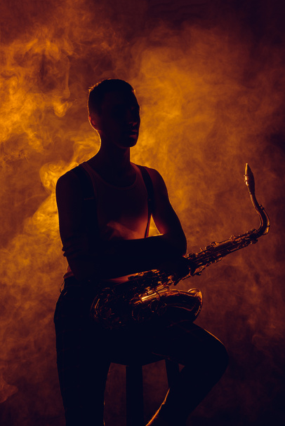 silueta de joven músico sentado en el taburete con saxofón en humo
 - Foto, imagen