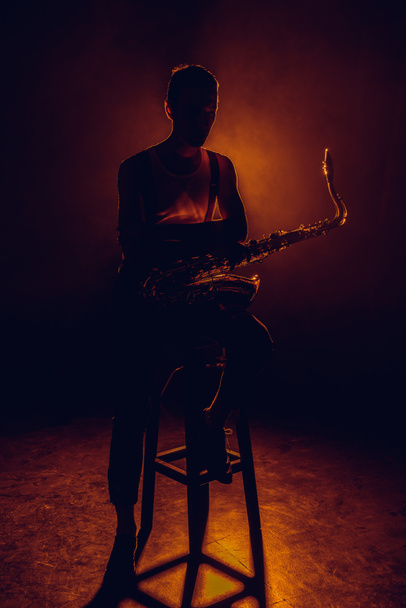 silueta de saxofonista joven sentado en el taburete con saxofón
  - Foto, Imagen