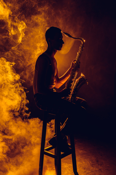 vista lateral del joven músico sentado en el taburete y sosteniendo el saxofón en el humo
  - Foto, imagen