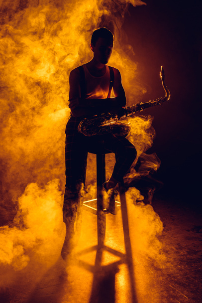 silueta del joven músico sentado en el taburete y sosteniendo el saxofón en el humo
  - Foto, Imagen
