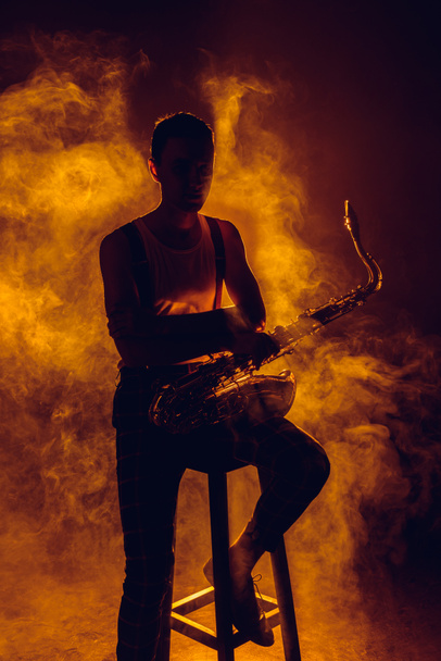 silhouette of young jazzman sitting on stool and holding saxophone in smoke  - Fotografie, Obrázek