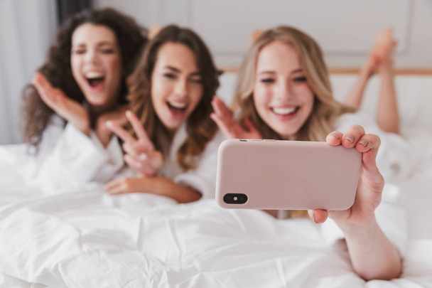 Blurry photo of happy smiling women 20s wearing white bathrobe lying in luxury bedroom in posh apartment and taking selfie on smartphone during hen party - Foto, afbeelding