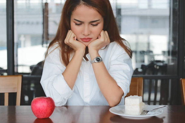 Mujer asiática molesto elegir entre manzana o pastel poco saludable en el escritorio. Estilo de vida saludable y concepto de dieta. Enfoque selectivo y profundidad de campo poco profunda
. - Foto, Imagen