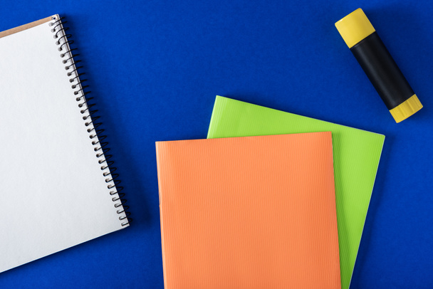 top view of blank notebook, glue stick and textbooks on blue background - Foto, afbeelding
