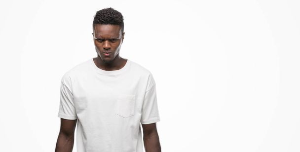 Joven hombre afroamericano vistiendo camiseta blanca escéptico y nervioso, frunciendo el ceño molesto debido a un problema. Persona negativa
. - Foto, Imagen