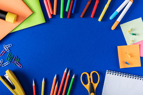вид сверху на красочные маркеры и карандаши, пустой учебник и разнообразные канцелярские принадлежности на синем фоне
 - Фото, изображение