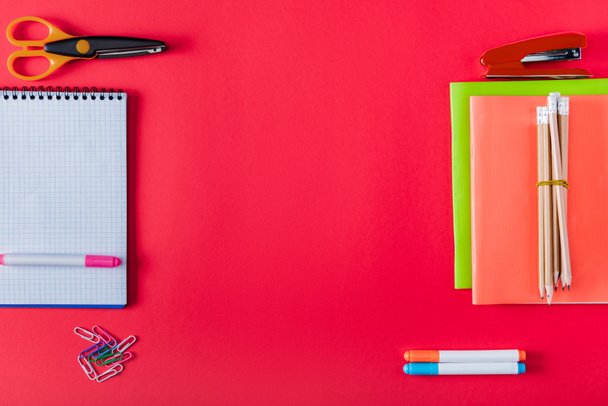 top view of arranged textbooks, stapler, markers, paper clips, scissors and blank notebook on red background  - Fotografie, Obrázek