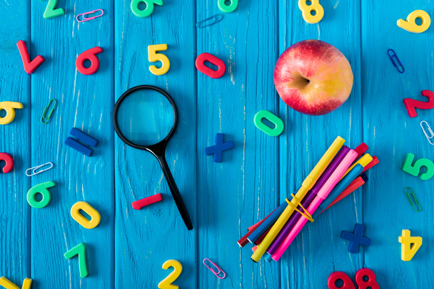 top view of magnifier, apple, colorful markers and various numbers on blue wooden background  - Fotoğraf, Görsel