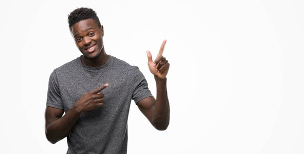 Jonge Afro-Amerikaanse man dragen grijs t-shirt glimlachen en kijken naar de camera met twee handen en vingers wijzen naar de kant. - Foto, afbeelding