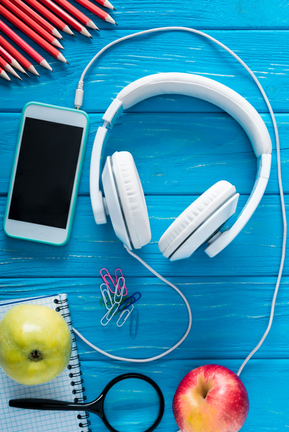 top view of headphones, smartphone with blank screen, apples and stationery on blue wooden background  - Fotoğraf, Görsel