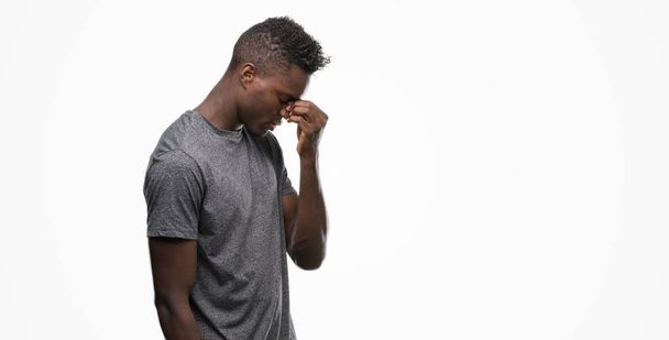 Молодой африканский американец в серой футболке устал тереть нос и глаза, чувствуя усталость и головную боль. Концепция стресса и разочарования
. - Фото, изображение