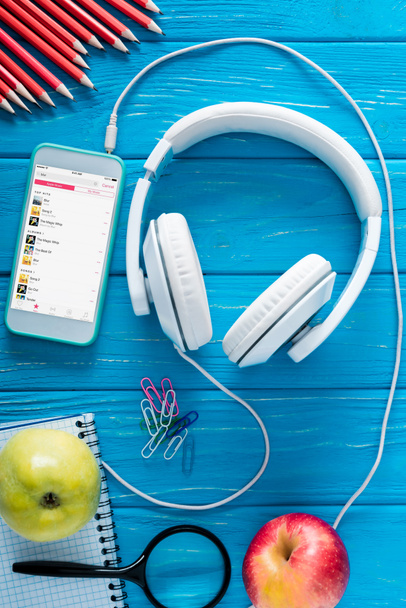 top view of smartphone with apple music application on screen, headphones, apples and stationery on blue wooden background  - Photo, Image