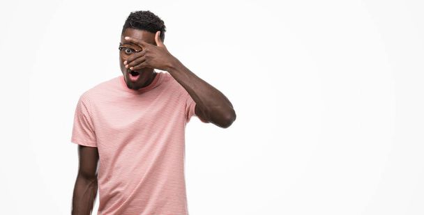 Jovem afro-americano vestindo camiseta rosa espreitando em choque cobrindo rosto e olhos com a mão, olhando através de dedos com expressão embaraçosa
. - Foto, Imagem
