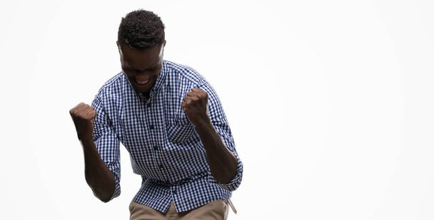 Young african american man wearing blue shirt very happy and excited doing winner gesture with arms raised, smiling and screaming for success. Celebration concept. - Foto, imagen