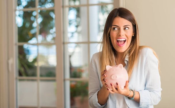 Beautiful young woman holding piggy bank at home, very happy with a smile because of profit. Business concept. - 写真・画像