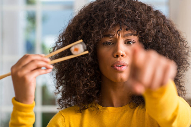 Donna afroamericana che mangia sushi con le bacchette a casa puntando il dito verso la macchina fotografica e verso di te, segno della mano, gesto positivo e fiducioso dal davanti
 - Foto, immagini