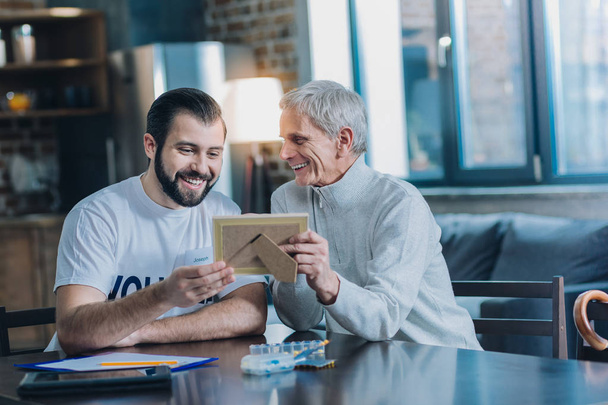 My family. Happy grey-haired man smiling and showing photos to a young man - Photo, Image