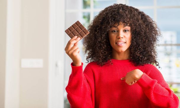 Африканская американка ест шоколадный батончик дома с удивленным лицом, указывающим пальцем на себя
 - Фото, изображение