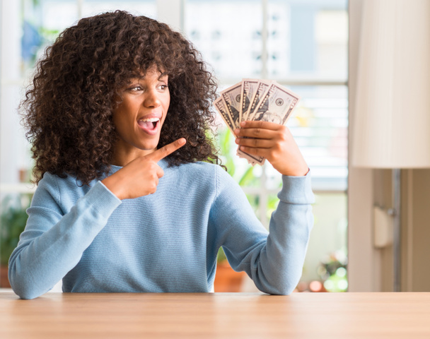 Mujer afroamericana sosteniendo billetes de banco de dólar muy feliz señalando con la mano y el dedo
 - Foto, imagen