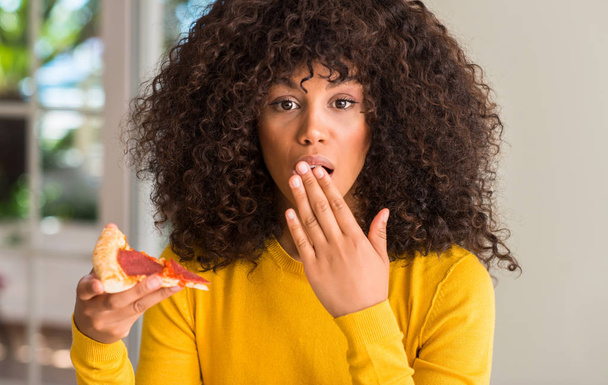 African american vrouw klaar om te eten peperoni pizza slice cover mond met de hand geschokt met schaamte voor fout, uitdrukking van angst, bang in stilte, geheime concept - Foto, afbeelding