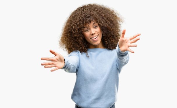 Donna afroamericana con un maglione che guarda la telecamera sorridente a braccia aperte per abbracciarsi. Espressione allegra che abbraccia la felicità
. - Foto, immagini