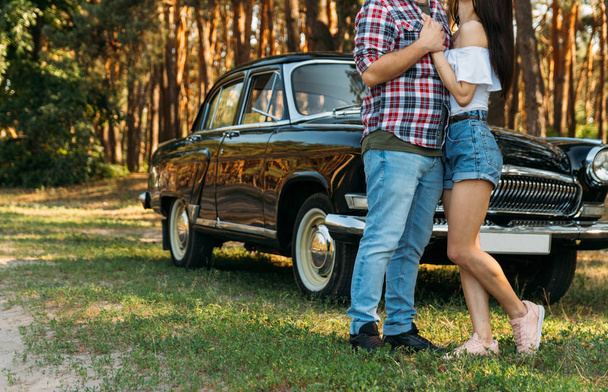 Love and affection between a young couple at the park. a guy in a plaid plane and jeans, a girl in shorts and a white jacket. They get together in the forest for a walk, near the old car. holding hands hugging - Photo, image