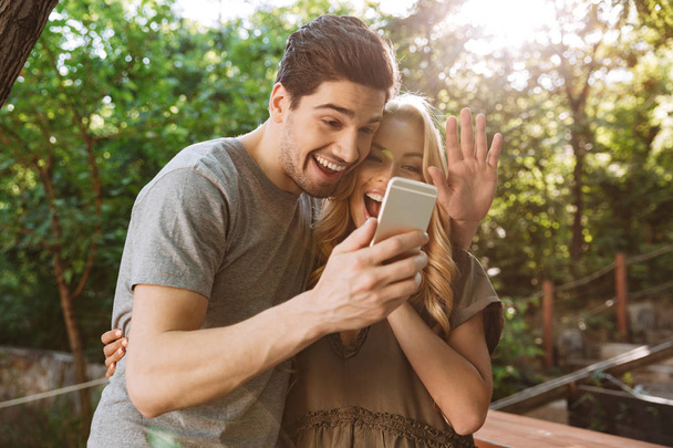 Фото улыбающейся молодой пары, позирующей вместе и делающей селфи на смартфоне на улице
 - Фото, изображение