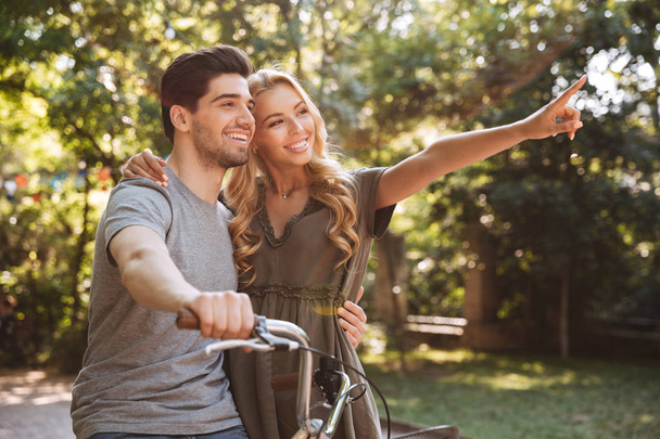 Picture of Pleased lovely young couple posing together with bicycle and looking away outdoors - Foto, Imagen