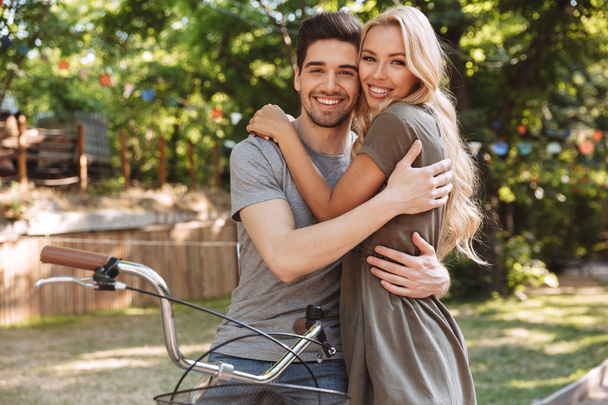 Smiling lovely young couple posing together with bicycle and looking at the camera outdoors - Фото, изображение