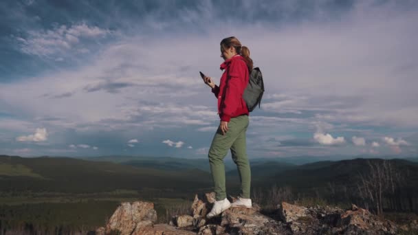 νεαρή γυναίκα ταξιδιώτη με σακίδιο και καπέλο κάνει μια selfie στο κινητό τηλέφωνο στέκεται στο χείλος του γκρεμού στα βουνά - Πλάνα, βίντεο