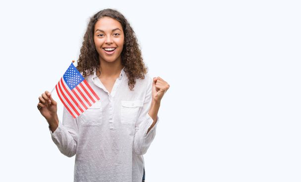 Young hispanic woman holding flag of United Estates of America screaming proud and celebrating victory and success very excited, cheering emotion - 写真・画像