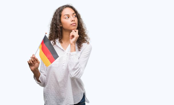 Giovane donna ispanica che tiene la bandiera della Germania faccia seria pensando alla domanda, idea molto confusa
 - Foto, immagini