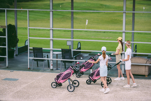 vista lateral de golfistas do sexo feminino com equipamento de golfe andando no campo de golfe no dia de verão
 - Foto, Imagem