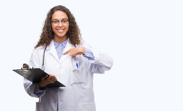 Молодая латиноамериканка-врач держит планшет с удивленным лицом, указывающим пальцем на себя
 - Фото, изображение