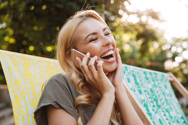 Side view of Cheerful blonde woman in dress sitting on sun lounger and talking by phone outdoors - Foto, afbeelding