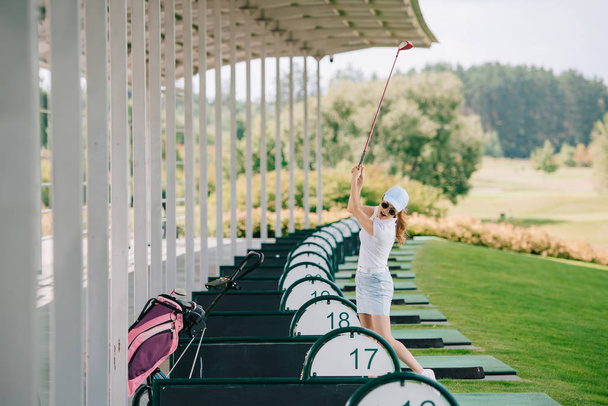 vrouw in polo, pet en zonnebril met golfclub in handen golfen op golfbaan - Foto, afbeelding