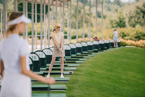 valikoiva painopiste hymyilevä naisten golfaaja poolossa ja korkki golfmailalla katsot kameraa golfkentällä
 - Valokuva, kuva
