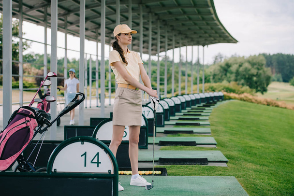 pensive female golfer in polo and cap with golf club looking away at golf course - Foto, imagen