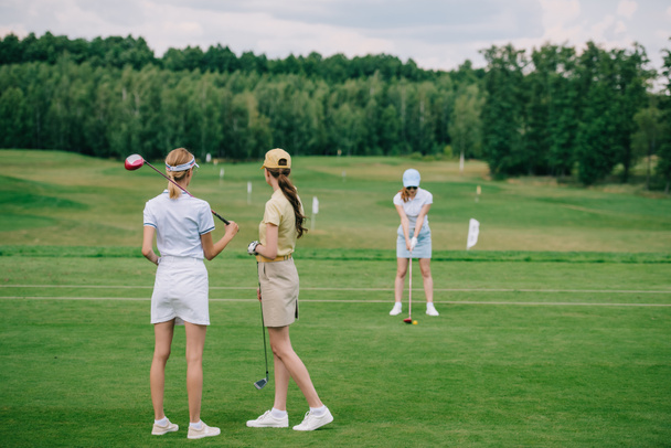 selective focus of women in caps with golf equipment looking at friend playing golf at golf course - Photo, Image
