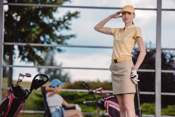 selective focus of female golf player playing golf while friend resting behind at golf course - Foto, afbeelding