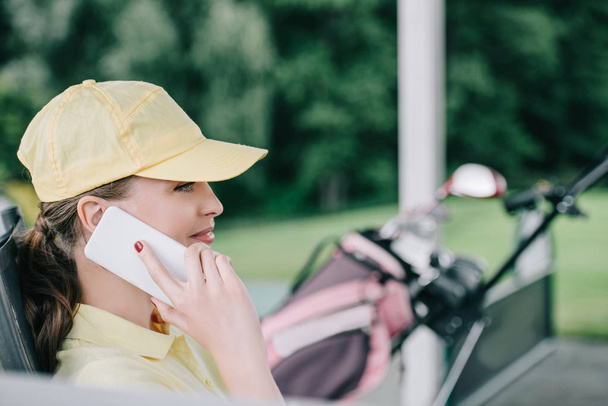 side view of female golf player in cap talking on smartphone at golf course - Photo, Image