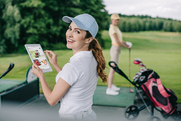 selective focus of smiling woman holding tablet with ebay logo and friend playing golf behind at golf course - Photo, image